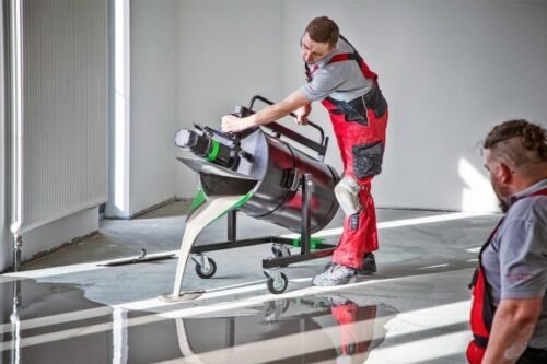 A worker using a cement mixer on a construction site, with another person observing in the background.